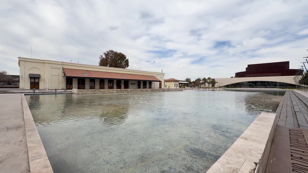 Panorámica lenta hacia la derecha sobre un estanque de agua poco profundo de hormigón en la Plaza del Bicentenario bajo cielos azules parcialmente nublados.