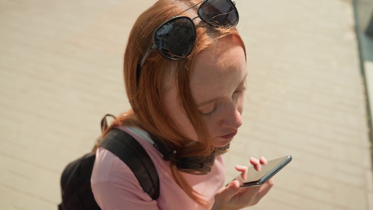 Giovane donna con capelli rossi parla al telefono mentre cammina all'aperto, indossando occhiali da sole e portando uno zaino. La donna occasionalmente guarda lo schermo del telefono durante la chiamata, mantenendo la concentrazione.
