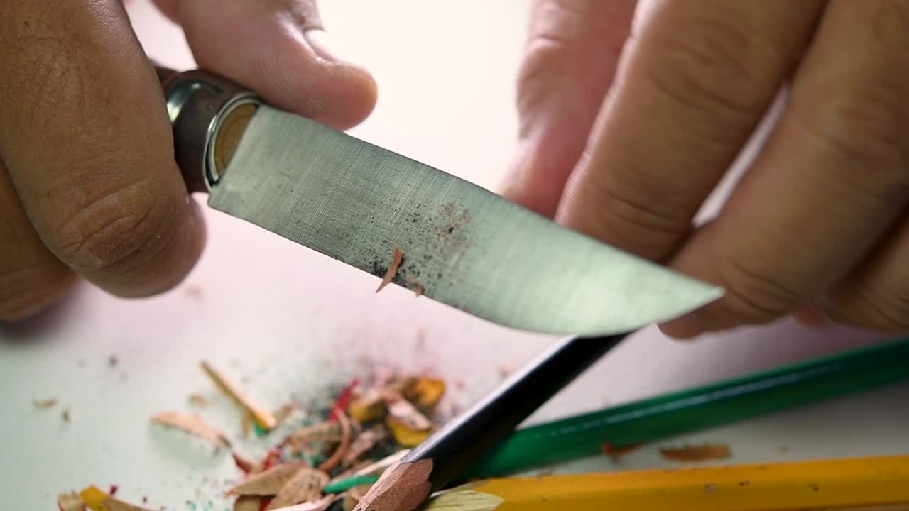 Footage of hands slowly sharpening a pencil and some coloured pencils with a sharp knife