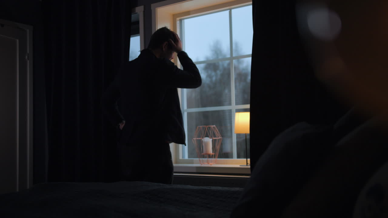 침실에서 시간을 확인하고 걱정스럽게 창 밖을 내다보는 남자의 정적인 모습
