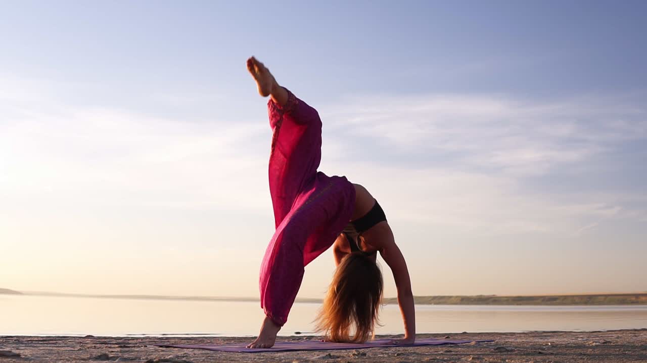Close up view of a young woman in pink yogi pants practicing on sand near the sea or lake in the morning. Posture - Urdhva dhanurasana