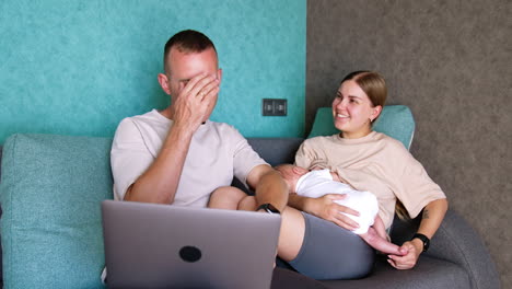 positive caucasian couple sit on the sofa. woman breastfeeding a newborn and man holds a laptop.