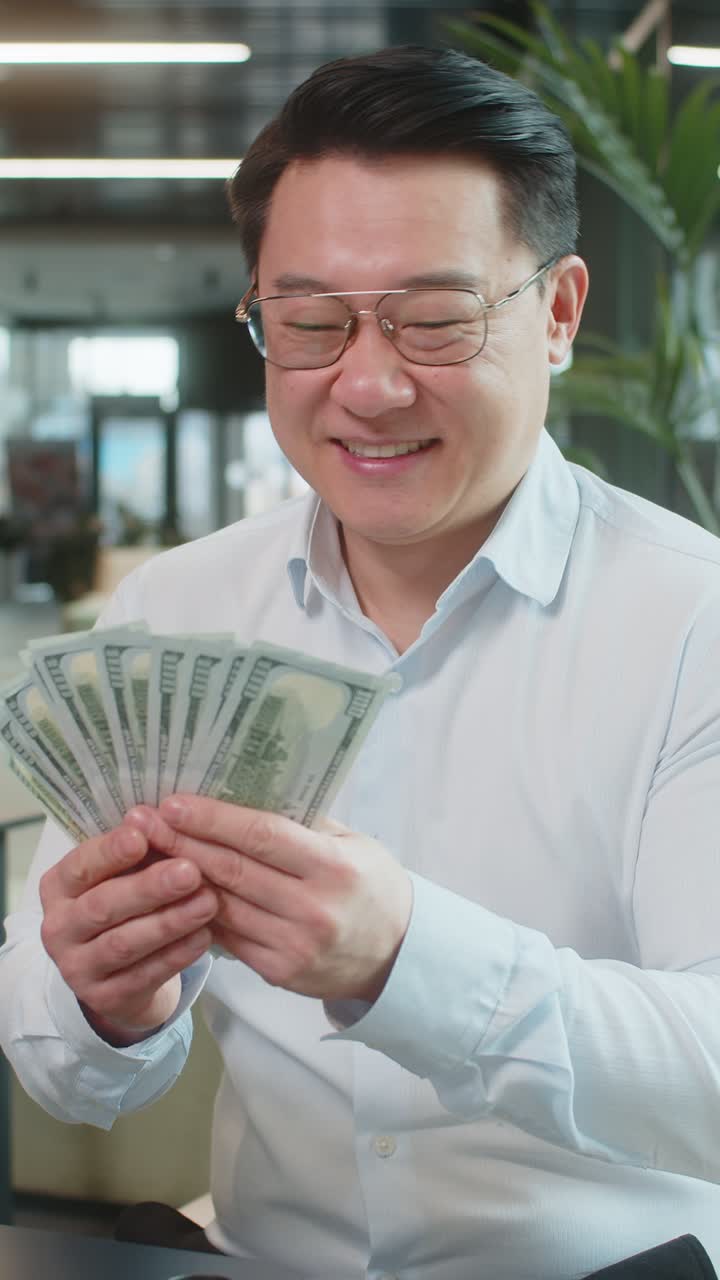 Smiling mature chinese businessman in formal shirt counting dollar banknotes in modern office lobby