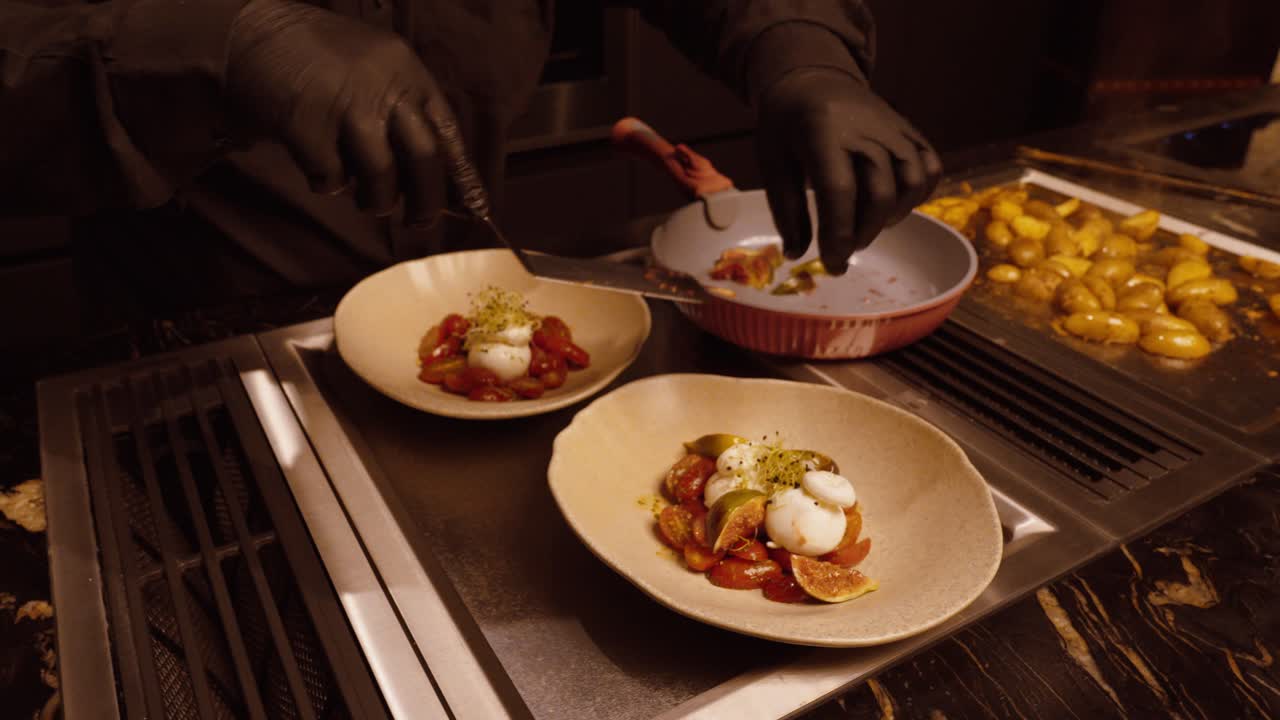 A chef wearing black gloves carefully arranges a gourmet dish with burrata and tomatoes in a modern kitchen, showcasing fine dining plating art