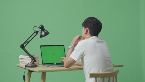 젊은 남성이 뭔가에 대해 생각하고 집에서 녹색 스크린 배경에서 녹색 화면 노트북에 타이핑하는 동안 주위를 둘러보고 있습니다.