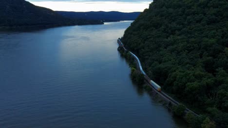 Drohnenaufnahmen-Aus-Der-Luft-Eines-Langen-Güterzuges,-Der-Bei-Sonnenuntergang-Entlang-Der-Gleise-Am-Fuße-Eines-Berges-Neben-Einem-Fluss-In-Den-Appalachen-Fährt