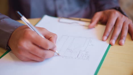person in long sleeve shirt drawing architectural sketch of hotel on white paper using black pen, hands resting on desk with phone and clip visible, suggesting creative focus and structural planning session