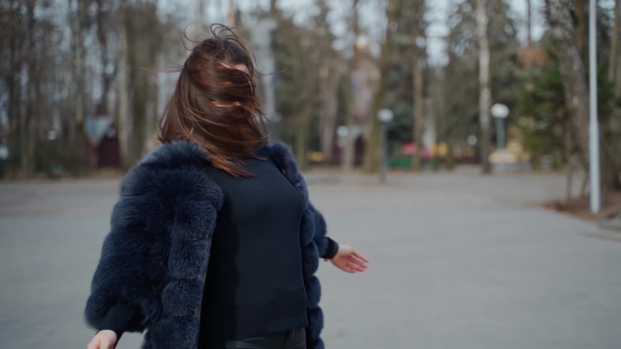 Pretty model in fur coat. Beautiful young woman is spinning happily in the park. Charming girl with long dark hair posing on camera outside.