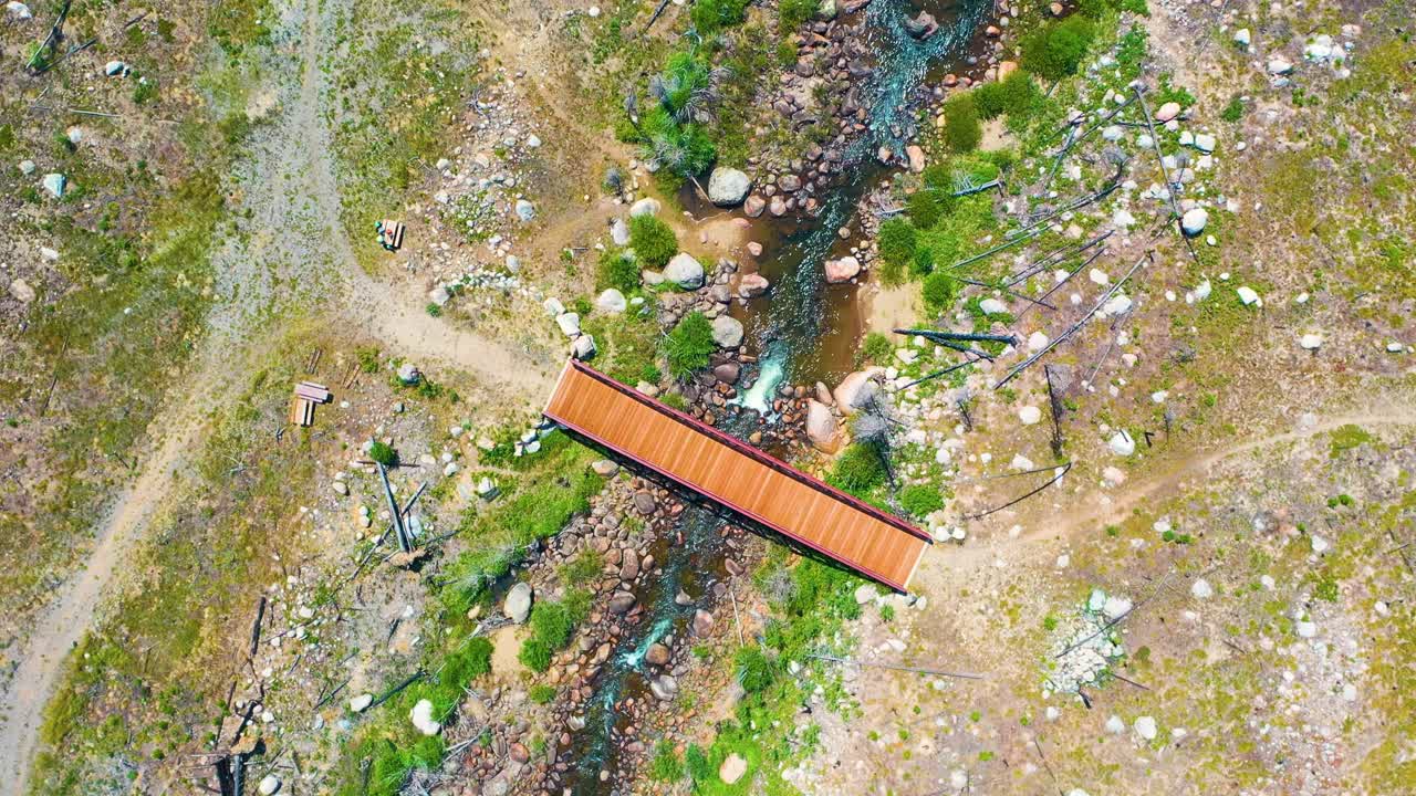 Riprese aeree verticali che seguono un ruscello tortuoso attraverso un terreno aspro e selvaggio, mentre un ponte di legno appare sopra il canale roccioso accanto a sentieri, vegetazione rada e terreno alpino
