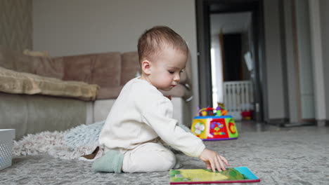 lovely toddler in white clothes sitting on the floor in the room. baby boy looking at book and eating a cookie.