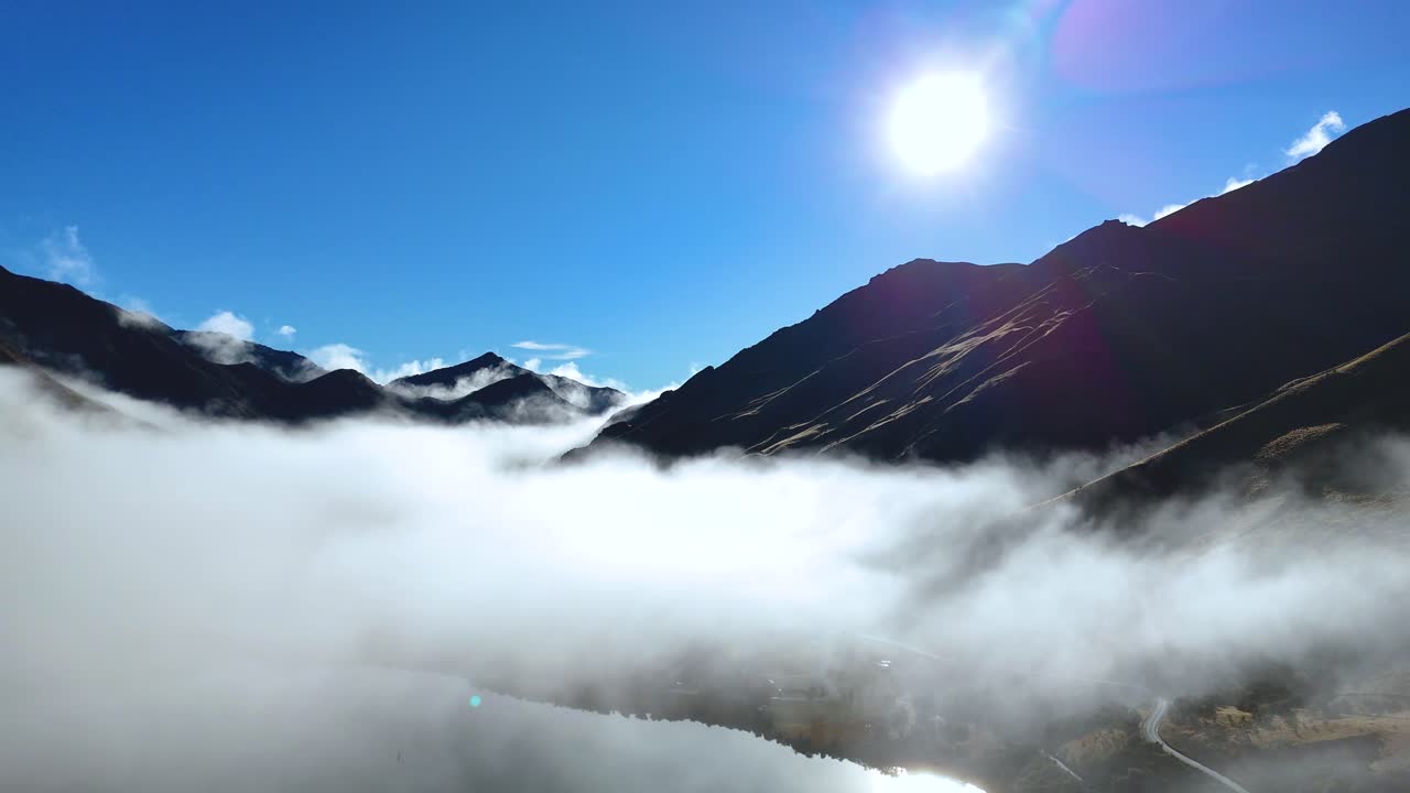 Fog drifts over Moke Lake against a backdrop of sunlit mountains in Queenstown, New Zealand. Bright, clear skies enhance the serene atmosphere