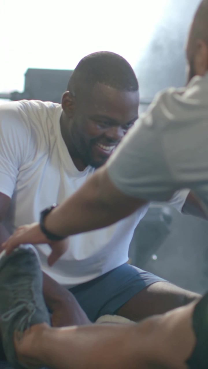 Vertical video: Camera pulling back assistant in gray tee reaching, stretching his leg on gym bench