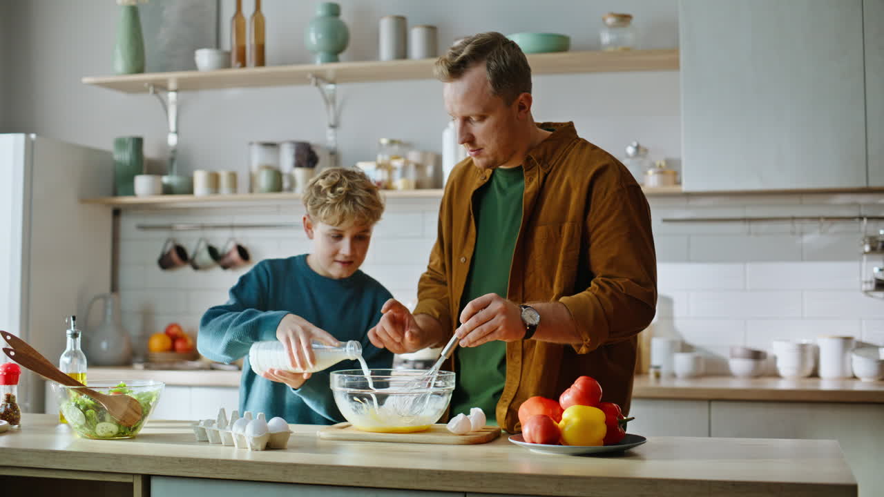 Nahaufnahme: Kleiner Sohn gibt Milch in Pfannkuchenteig und bereitet mit seinem Vater das Frühstück zu.