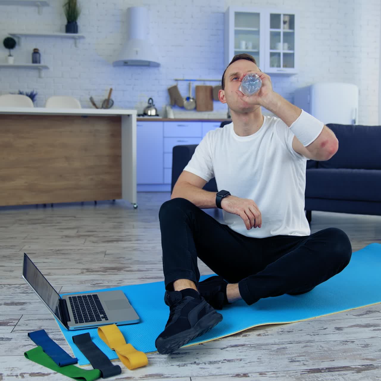 Man drinks water after daily workout at home. Guy is sitting on a fitness mat next to the laptop and holding a bottle of water. Fitness indoors. Sport during quarantine