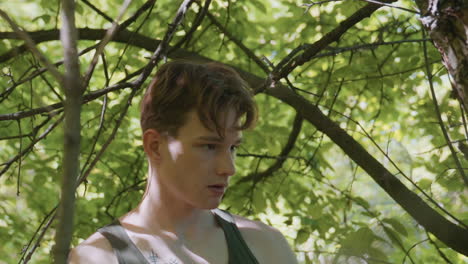 young man unties headband while scanning surroundings with intense gaze and careful movements beneath leafy branches and thorns near rugged tree trunk, sunlight dappling muscular arms