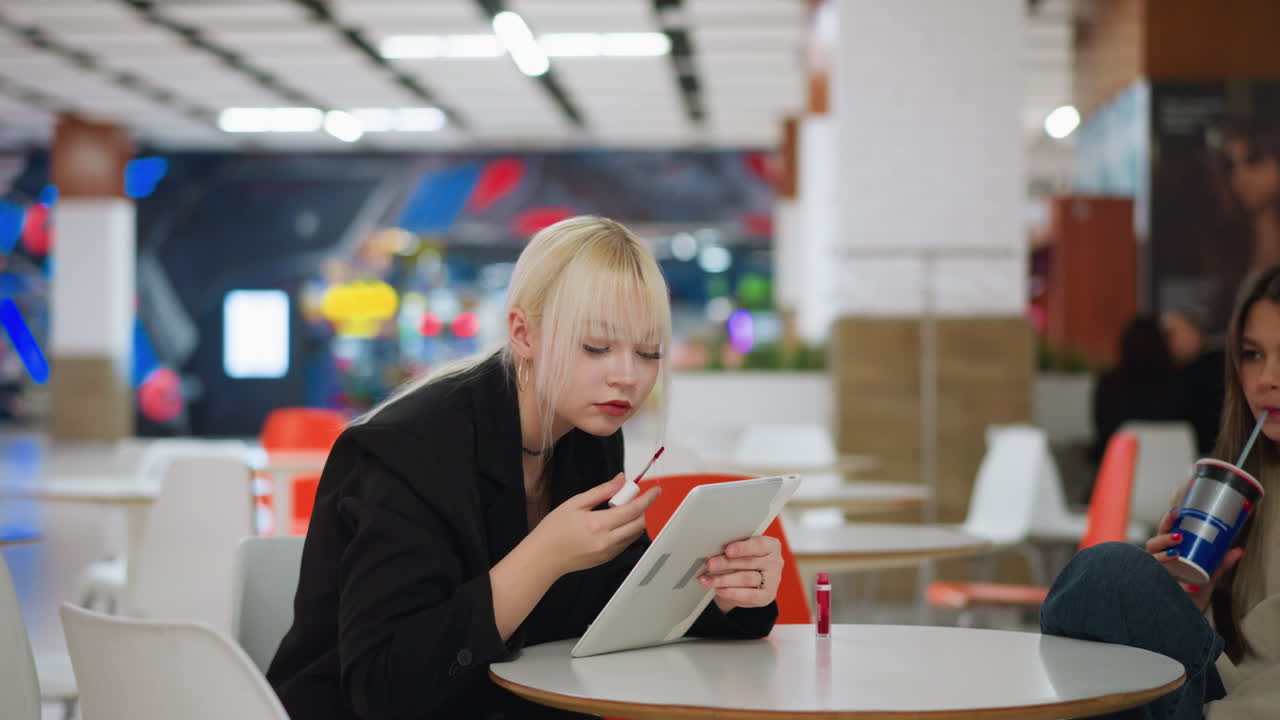 Woman applying lipstick using tablet mirror while seated in cafe, friend beside sipping drink with straw, showing focus on beauty routine and casual lifestyle indoors with blurred colorful background