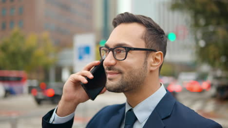 Porträt-Eines-Glücklichen-Geschäftsmannes,-Der-An-Einer-Belebten-Straße-In-Der-Stadt-Steht-Und-Mit-Seinem-Mobiltelefon-Telefoniert,-Während-Der-Verkehr-Im-Hintergrund-Verschwommen-Ist