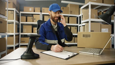 Young-Man-In-Uniform-Talking-On-Mobile-Phone-While-Working-At-Laptop-Computer-In-Post-Office-Store-With-Parcels