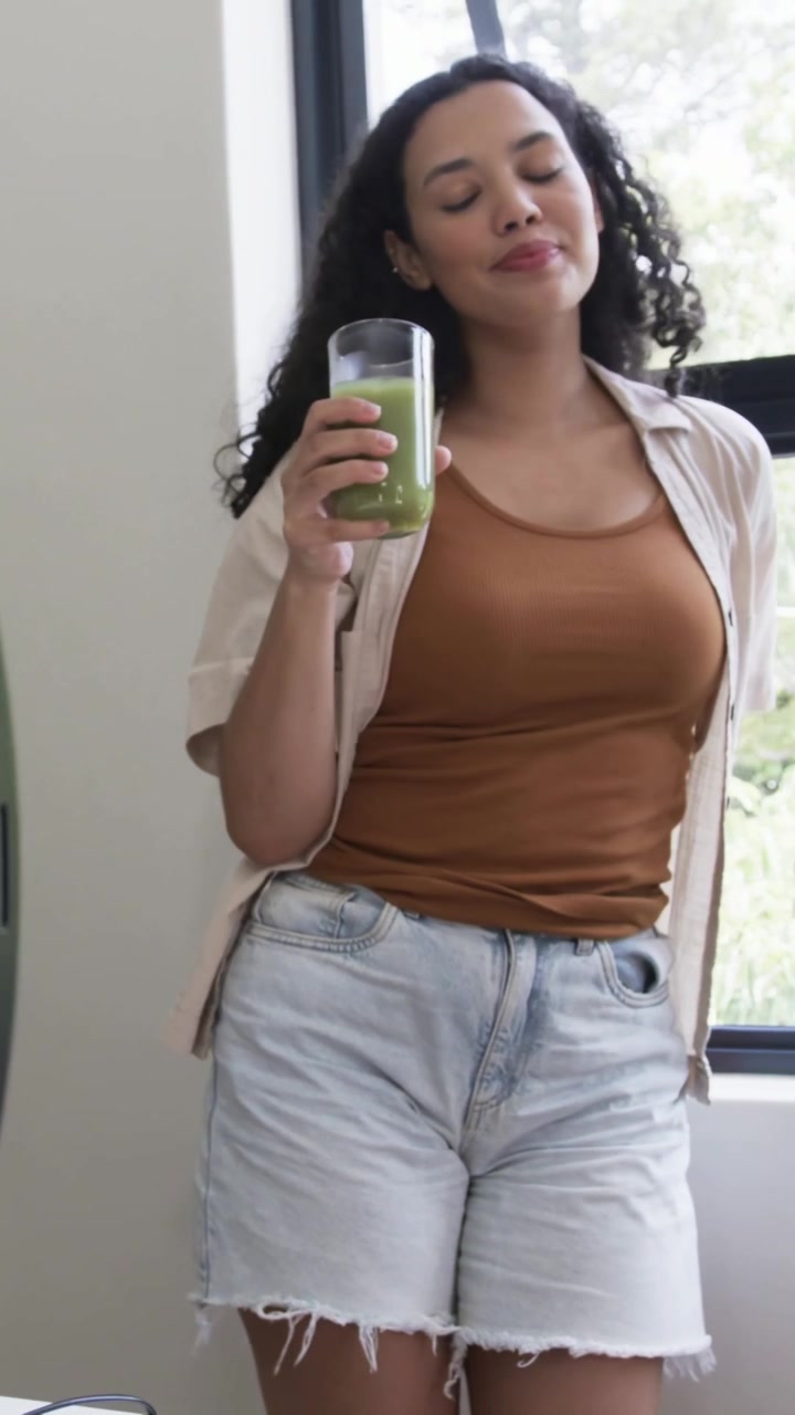 Video verticale: Una modella in top sorseggia succo verde vicino a una finestra, con la telecamera che panoramica, mentre prepara un ring light.