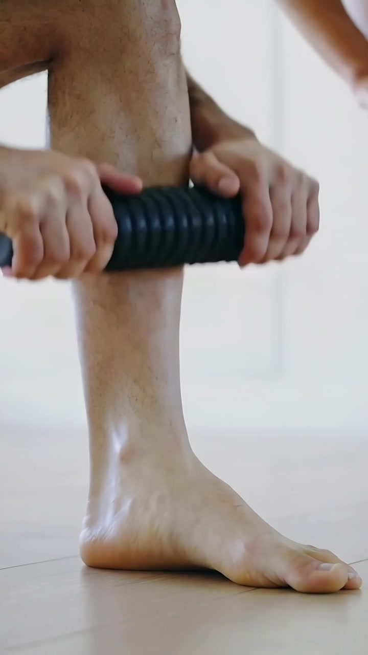Vertical video: Rolling man in gym shorts rubbing calf with roller at home after workout for relief
