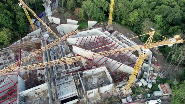 Luftaufnahme: Bau eines neuen Gebäudes mit hohen Turmkränen in grüner Umgebung.