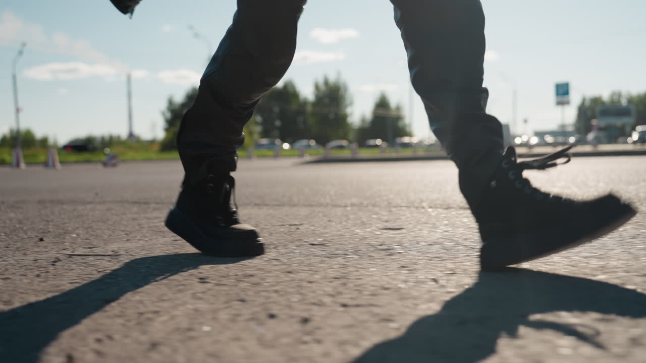 Close up of black boots walking on asphalt street casting long shadow in bright sunlight with blurred cars, trees, and city background symbolizing strength, determination, urban lifestyle