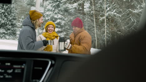 cheerful friends drinking hot tea in a snowy forest during a winter road trip