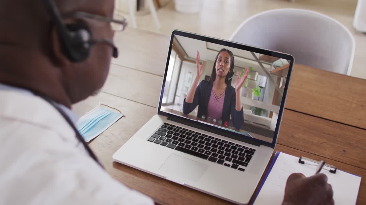 Afroamerikansk mandlig læge med headset på, der tager noter under et videoopkald på sin bærbare computer