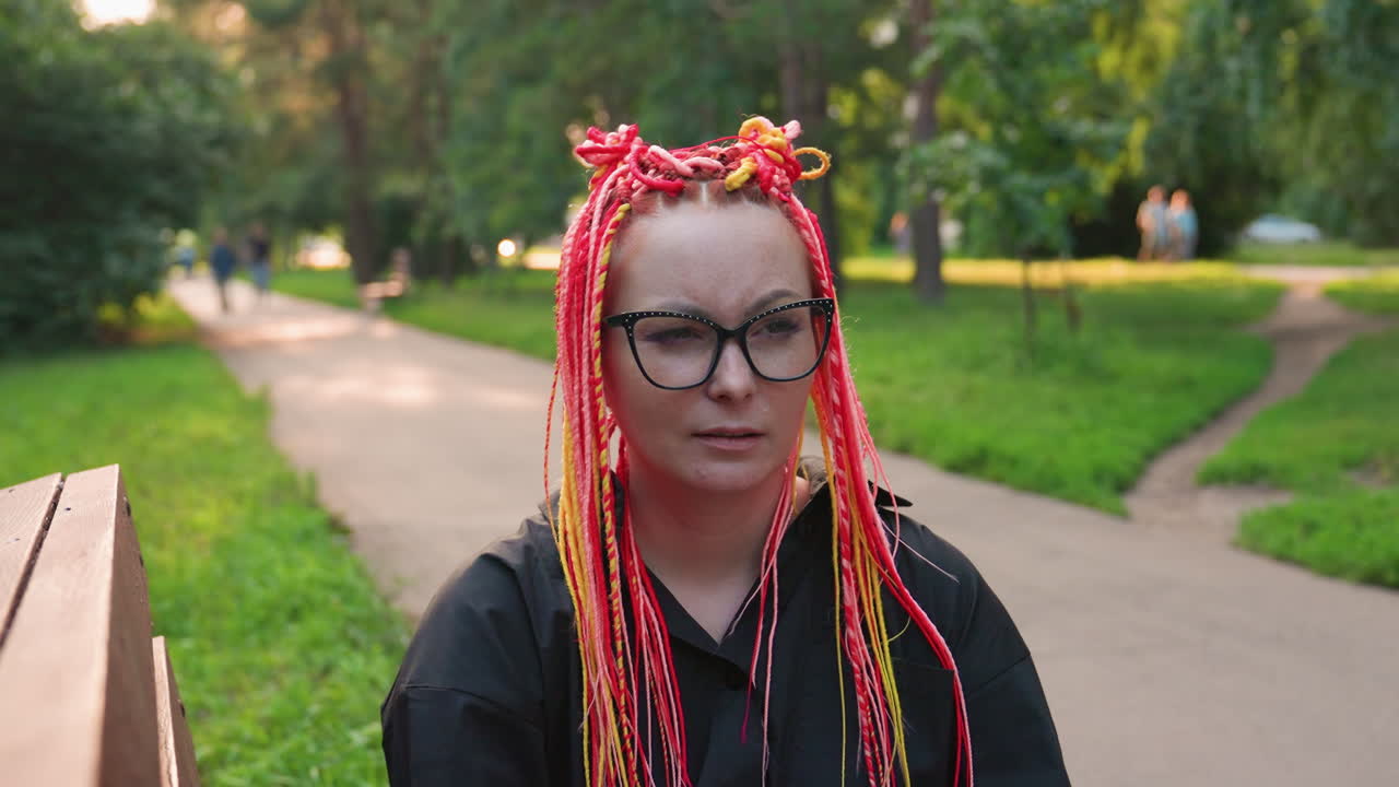 Una joven blanca con trenzas coloridas se sienta en un banco del parque, mira hacia el sendero arbolado, se ajusta las gafas, se detiene durante un paseo tranquilo y muestra una tranquila tristeza e introspección, lleva una sudadera negra suave.
