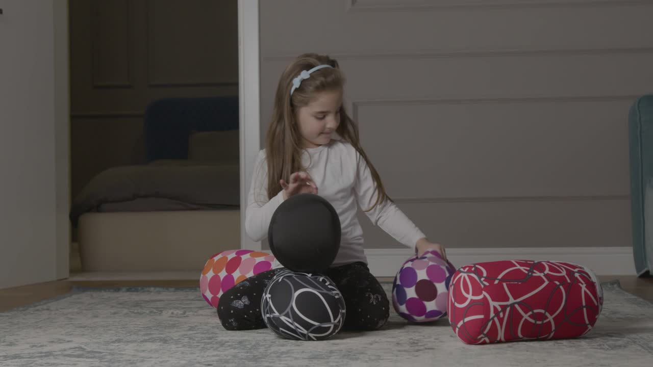 Una bambina che tenta di impilare cuscini rotondi sul pavimento prima che cadano.