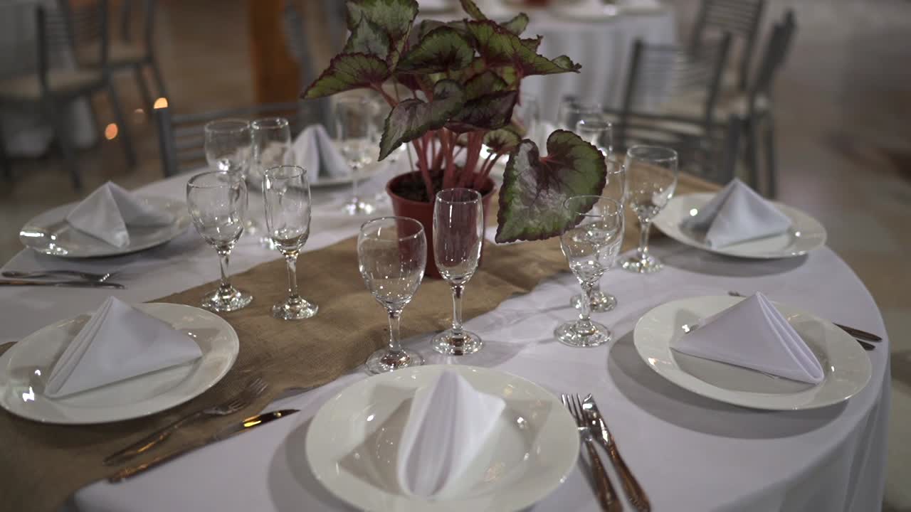 Elegant round wedding reception table features white place settings with folded napkins glassware a potted begonia centerpiece and burlap runner
