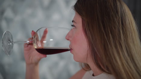 close up side view of young woman drinking red wine quickly from glass, wearing sleeveless top, long hair flowing down, indoor setting with softly lit geometric background