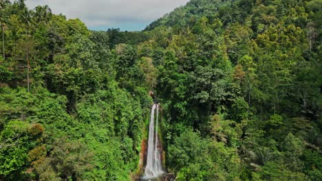 a stunning aerial drone shot of gitgit waterfall in bali, flowing in full view, surrounded by tropical trees and vibrant green foliage, with clouds and blue sky visible above