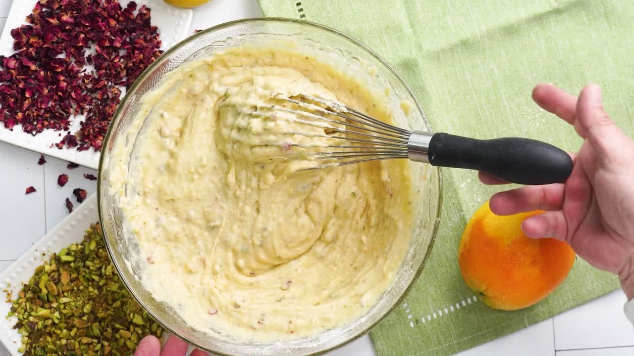 Baking a dessert called Persian Love Cake made with rose petals and pistachios. Cake with white glaze frosting. Using a whisk to combine ingredients. Beautiful cake for tea time or celebration