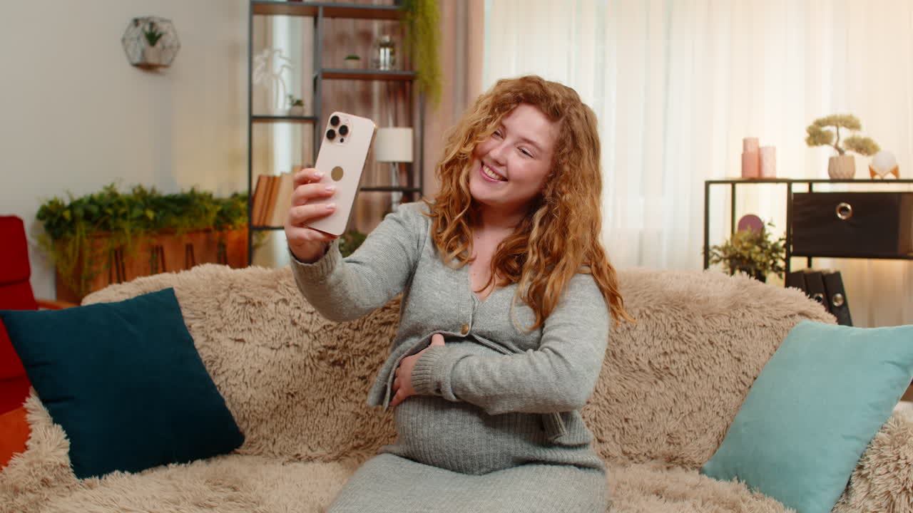 Pregnant young woman talking on video call with friend on smartphone showing belly smiling with joy