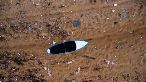 aerial top down, person holding surfboard above head walking on trash polluted beach