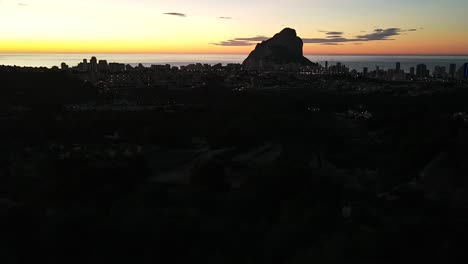 Drohnenaufnahmen,-Die-Eine-Atemberaubende-Silhouette-Der-Stadt-Calpe,-Spanien,-Und-Das-Naturdenkmal-Peñón-De-Ifach-Vor-Dem-Hintergrund-Eines-Wunderschönen-Gelben-Und-Orangefarbenen-Sonnenaufgangs-An-Einem-Frühen-Sommermorgen-Zeigen