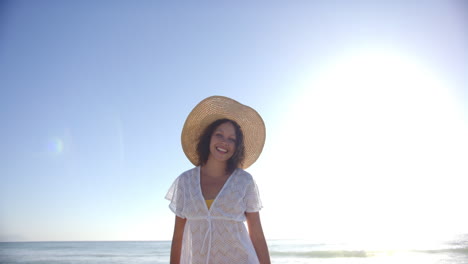 Una-Joven-Birracial-Se-Encuentra-En-La-Playa,-Con-Un-Sombrero-De-Ala-Ancha-Y-Una-Bata-Blanca.