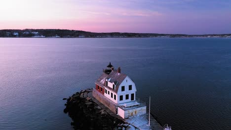 Low-aerial-ORBIT-around-a-white-building-attached-to-a-red-brick-lighthouse-at-the-end-of-a-snow-dusted-rocky-breakwater