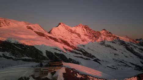 aerial-view-of-building-on-top-of-the-alps,-sunrise,-orange-lit-rocky-mountains