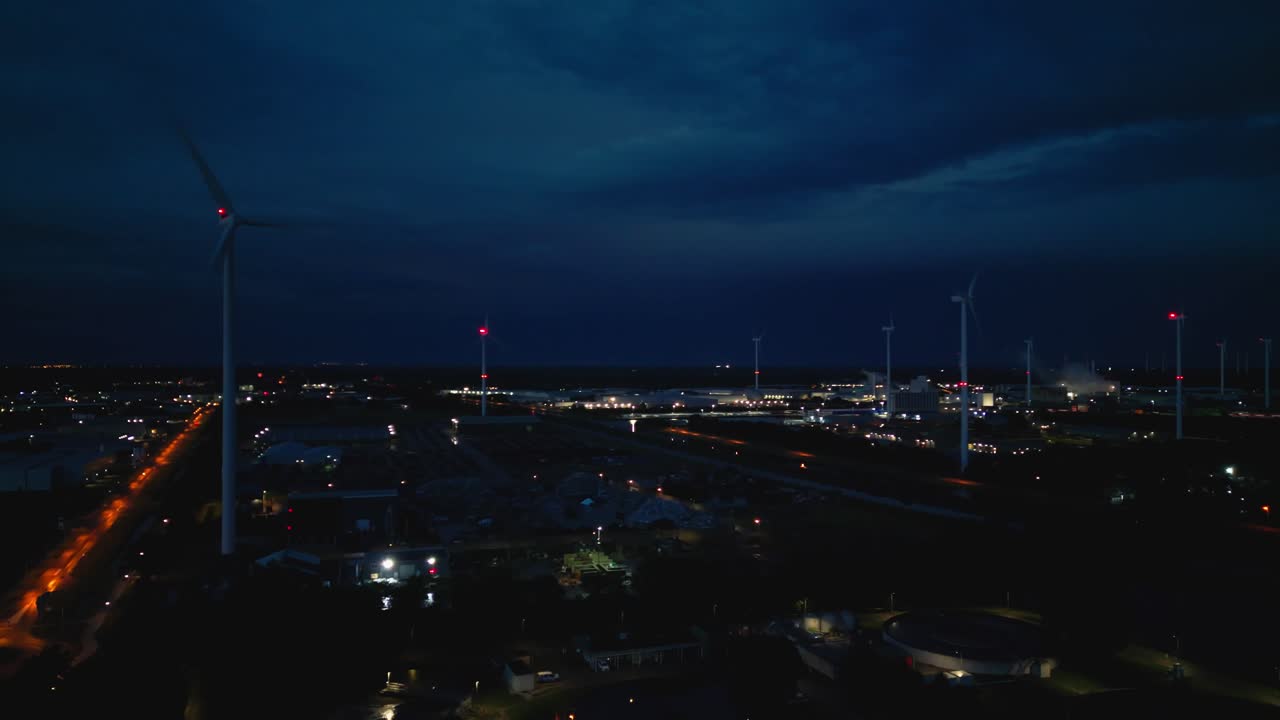 Drone footage of a wind turbine at night with glowing city lights, illuminated roads, and industrial facilities under a dark sky. Location: Coevorden, Drenthe, Netherlands