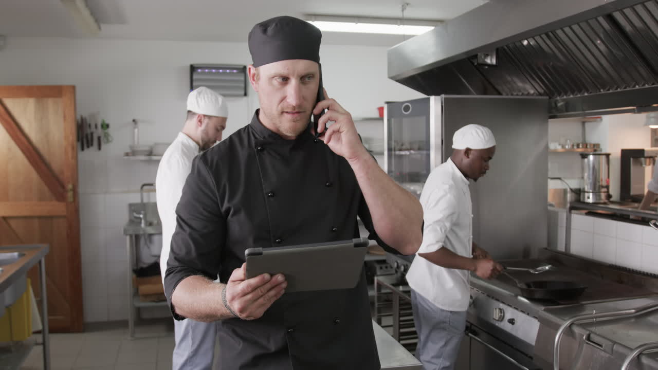chef caucásico usando tableta y hablando en teléfono inteligente en la cocina, cámara lenta