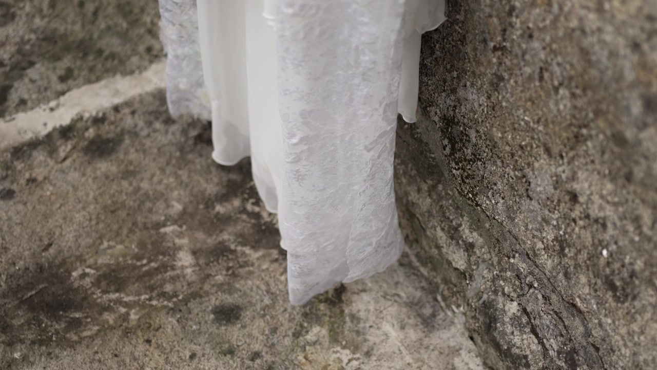 Close up of a wedding dress hem with intricate lace details against a stone wall