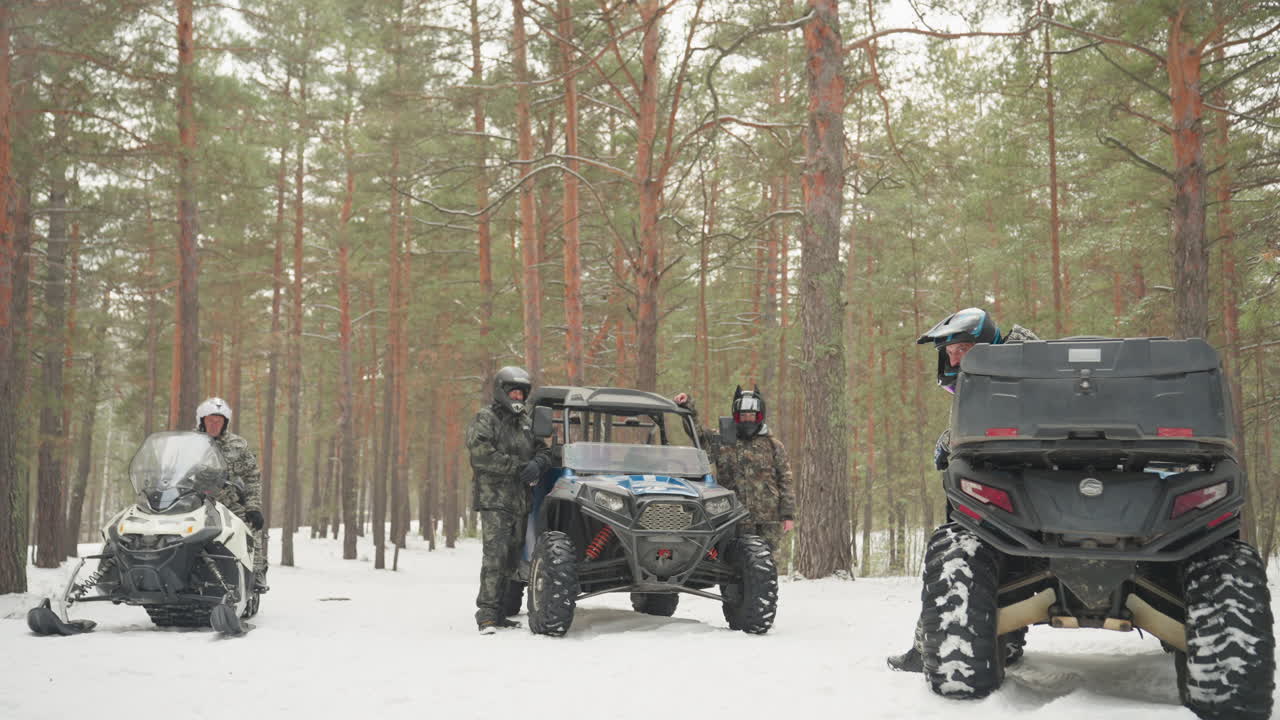 Un gruppo di piloti prepara un convoglio di quad in una foresta innevata, UTV e motoslitte parcheggiati allineati tra alti pini, l'equipaggio ispeziona attrezzatura e pneumatici prima della corsa fuoristrada, luce invernale calma e aria frizzante creano un'atmosfera concentrata.