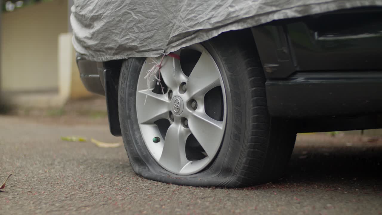 Flat car tire under a cover on an asphalt road showing a deflated rubber sidewall and alloy rim