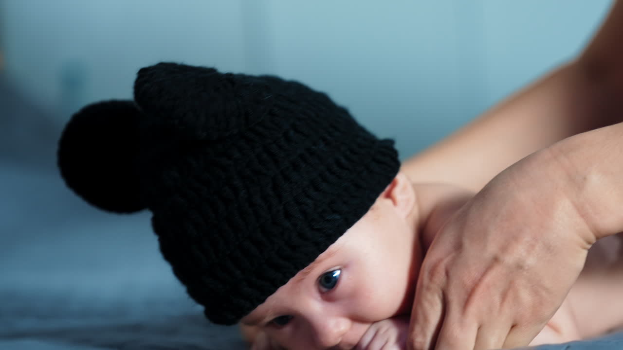 Sweet little baby in funny black cap lies face down. Parent's hands try to lift him but babe is busy with chewing his fist. Close up.