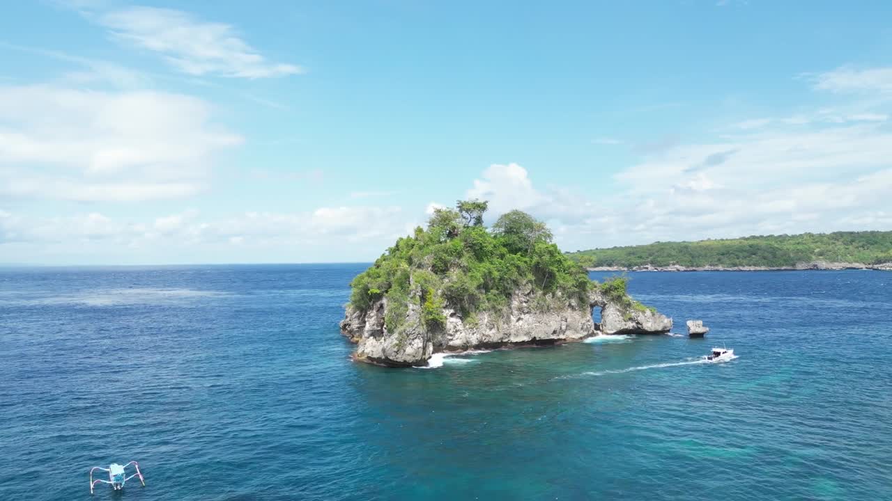Drone video of a tranquil Bali island cove, vivid blue waters, lush greenery, bright skies, and boats passing. Captivating aerial footage showcasing stunning tropical paradise