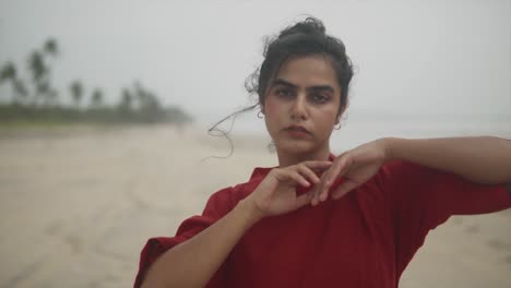 una joven morena encantadora en un vestido rojo se encuentra en una playa vacía, mirando hacia adelante y gestando con sus manos - primer plano
