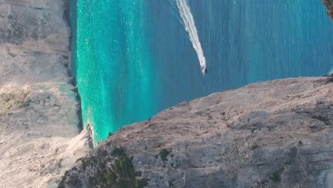 Vertical-Drone-Shot,-Motor-Boat-Sailing-in-Turquoise-Sea-Water-Under-Cliffs-of-Greek-Island-of-Zakynthos