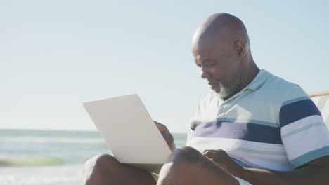 Happy-senior-african-american-man-sitting-on-deck-chair-and-using-laptop-at-beach,-in-slow-motion